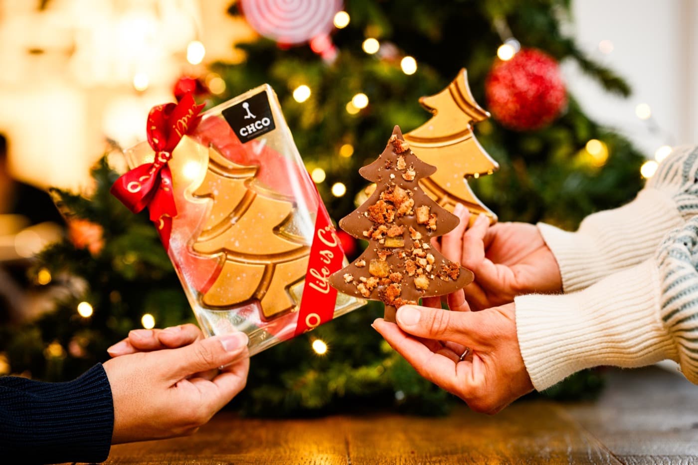 Twee handen met chocolade kerstbomen voor een versierde kerstboom